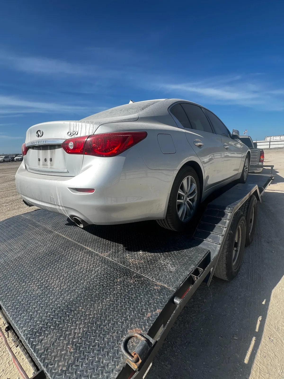 Rear three-quarter view of the dusty silver Q50 on the trailer showing streaked dirt down the trunk and bumper