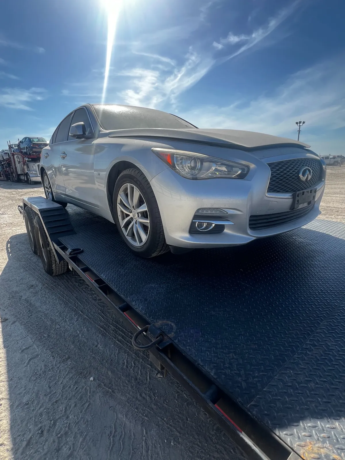 Dirty silver 2015 Infiniti Q50 strapped to a black car trailer on an auction lot